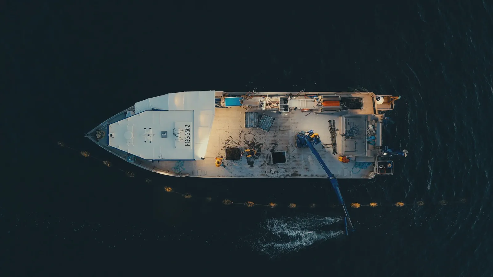 Vue aérienne du pont d’un bateau chargé de caisses et d’équipement, tuyau plongeant vers des bouées en mer.