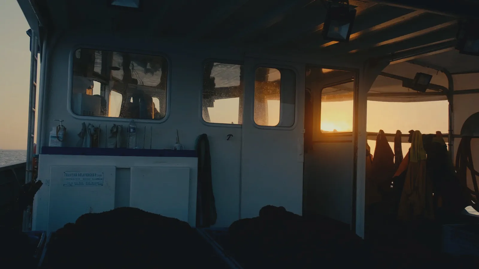 Intérieur d’une cabine de bateau au coucher du soleil, cirés suspendus, porte ouverte sur la lumière du soir.