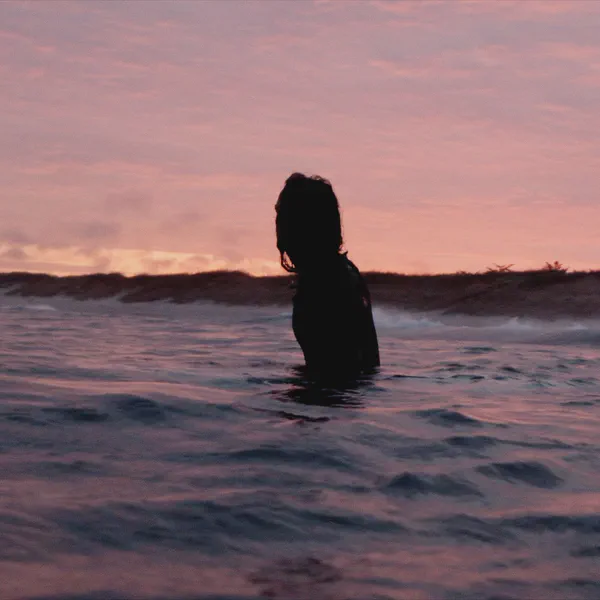 Silhouette d’une personne debout dans la mer jusqu’à la poitrine, ciel rose au coucher du soleil, dunes au loin.
