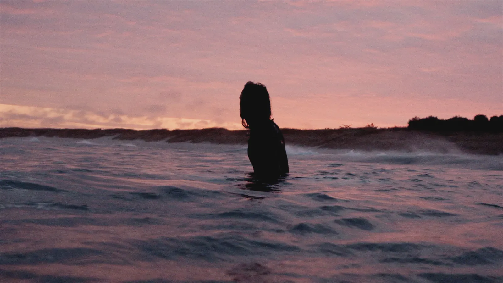 Silhouette d’une personne debout dans la mer jusqu’à la poitrine, ciel rose au coucher du soleil, dunes au loin.