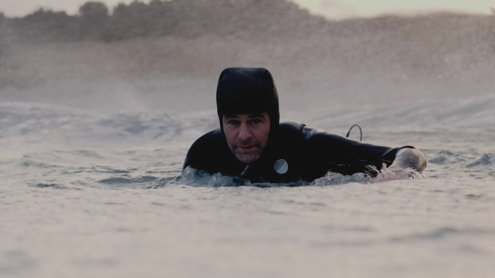 Yoann Renambot flotte dans l’eau, capuche de combinaison sur la tête, dunes floues en arrière-plan.
