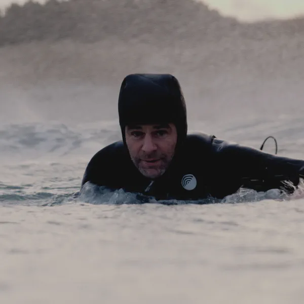 Yoann Renambot flotte dans l’eau, capuche de combinaison sur la tête, dunes floues en arrière-plan.
