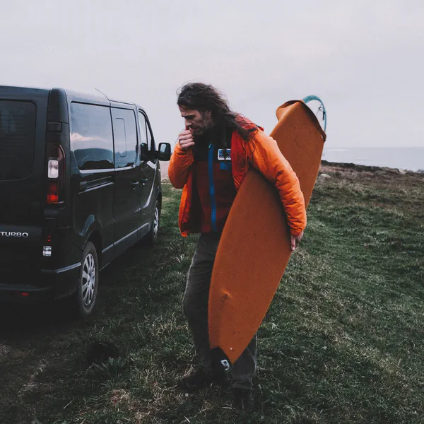 Yoann Renambot marche près d’un fourgon noir, planche de surf orange sous le bras, souffle visible dans l’air froid.