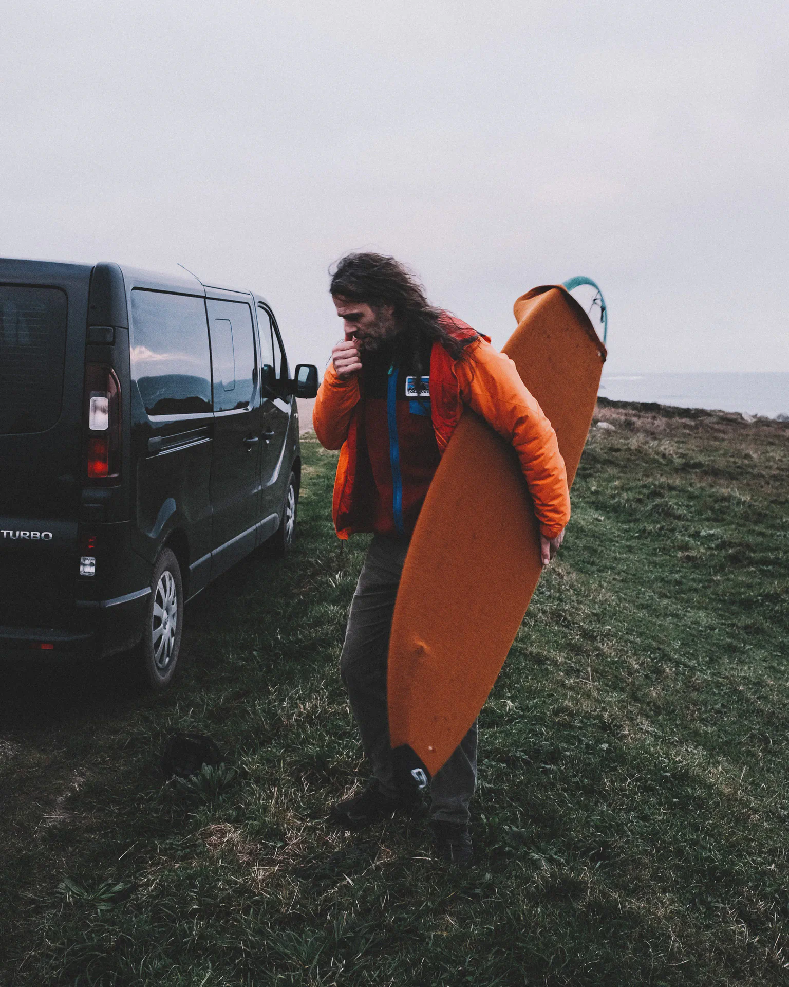 Yoann Renambot marche près d’un fourgon noir, planche de surf orange sous le bras, souffle visible dans l’air froid.