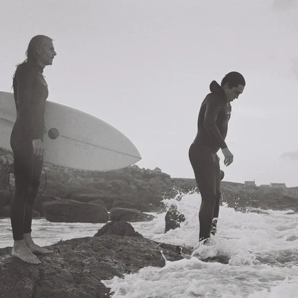 Deux surfeurs sur des rochers en bord de mer, l’un tenant deux planches, l’autre entrant dans une vague, noir et blanc.