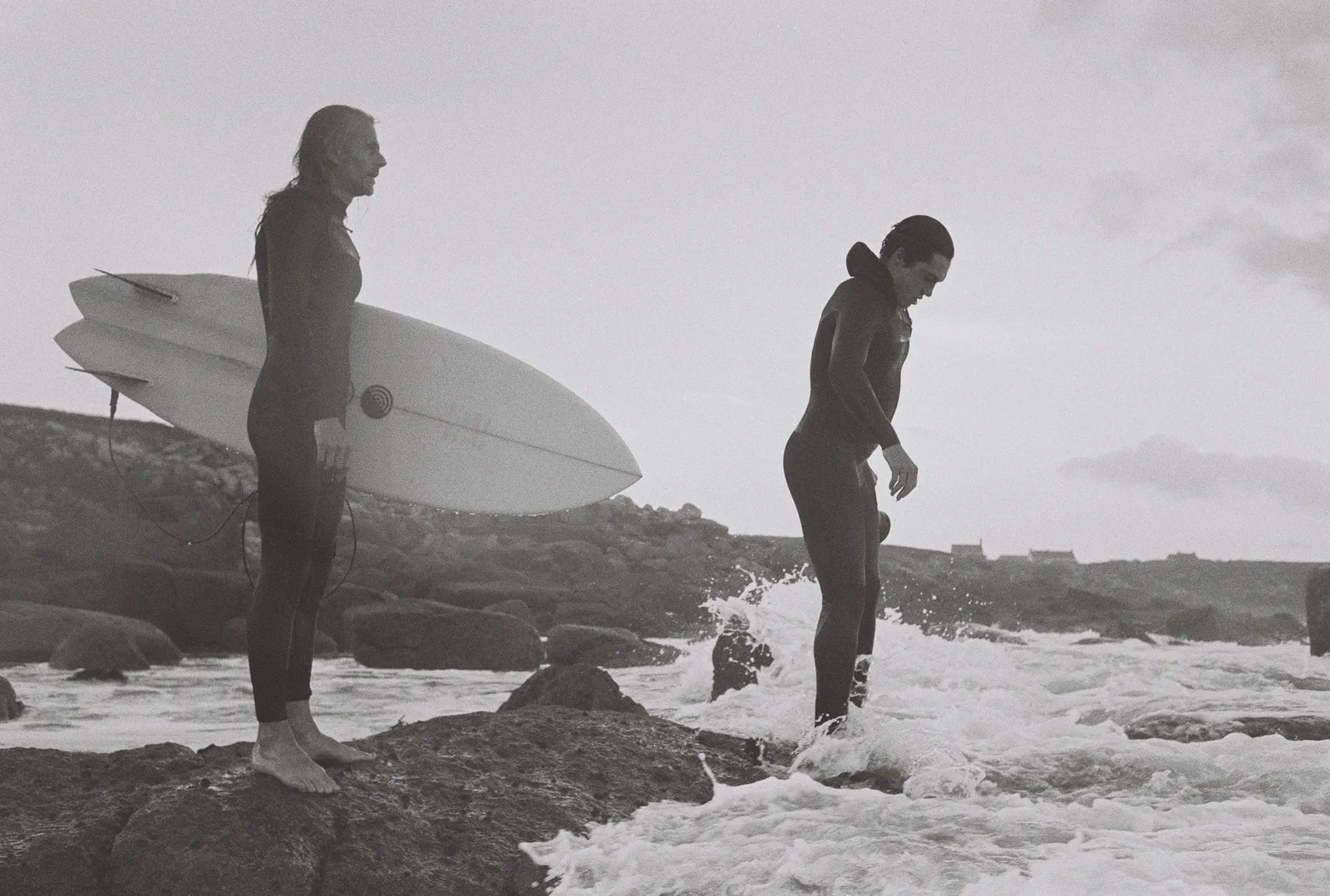 Deux surfeurs sur des rochers en bord de mer, l’un tenant deux planches, l’autre entrant dans une vague, noir et blanc.