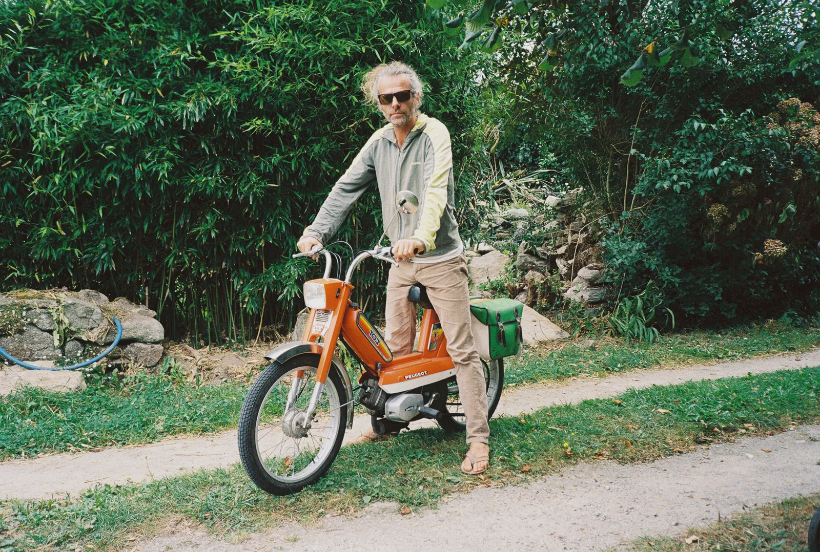 Un homme âgé en lunettes de soleil conduit un vieux cyclomoteur orange sur un chemin bordé de bambous.