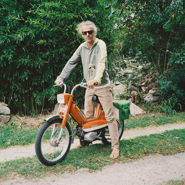 Un homme âgé en lunettes de soleil conduit un vieux cyclomoteur orange sur un chemin bordé de bambous.