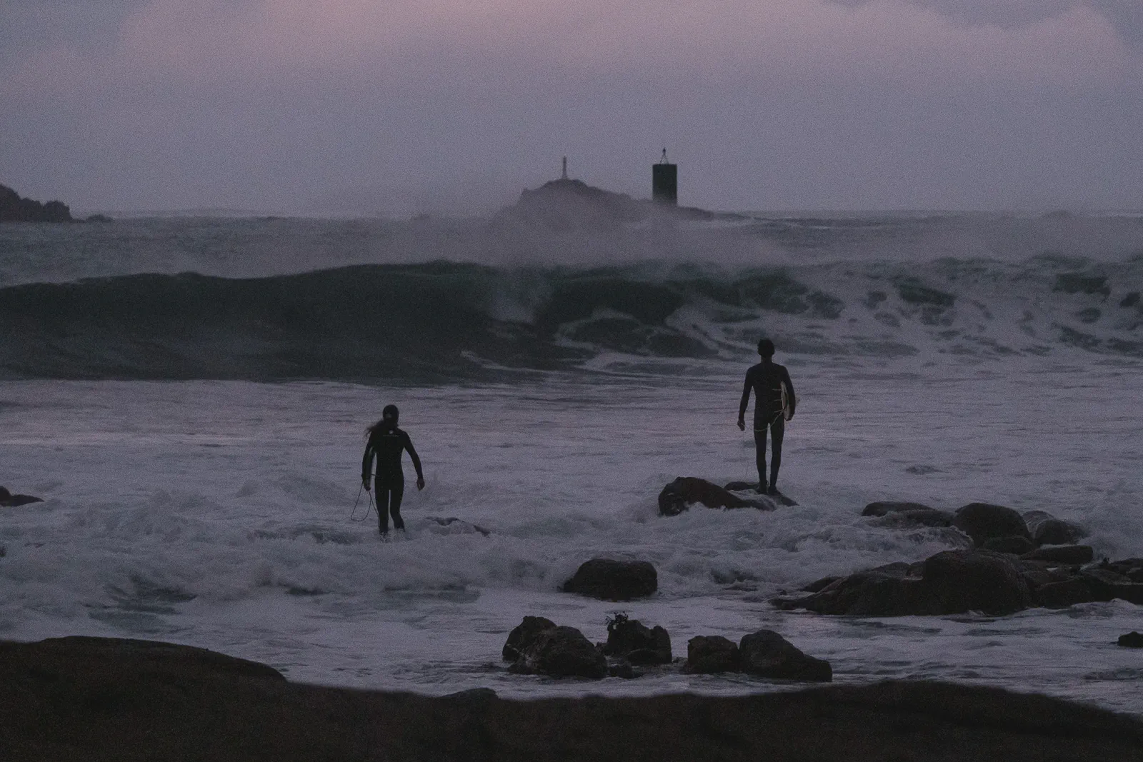 Deux surfeurs entrent dans une mer agitée et brumeuse au crépuscule, une balise de navigation visible entre les vagues.