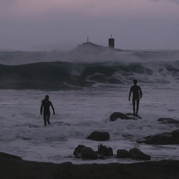 Deux surfeurs entrent dans une mer agitée et brumeuse au crépuscule, une balise de navigation visible entre les vagues.