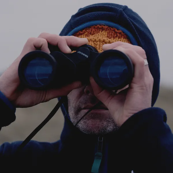 Un homme capuche observe l’horizon aux jumelles, bonnet orange visible sous la capuche, arrière-plan flou.