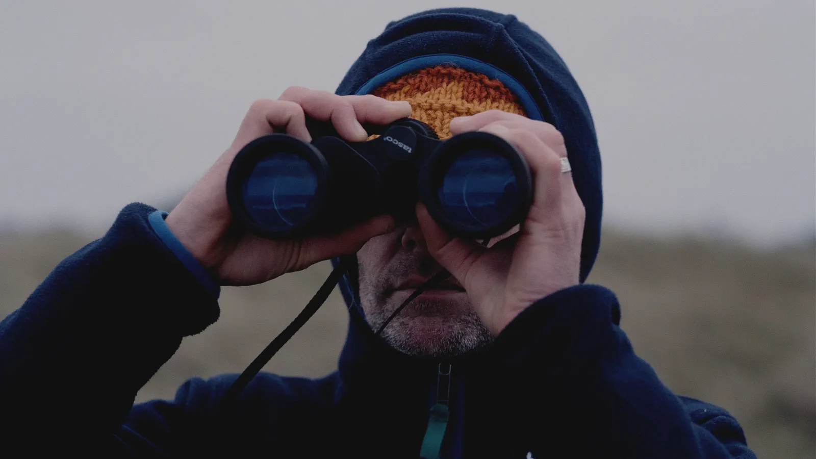 Un homme capuche observe l’horizon aux jumelles, bonnet orange visible sous la capuche, arrière-plan flou.