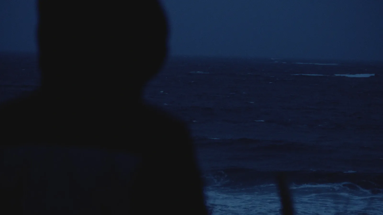 Silhouette d’une personne encapuchonnée face à une mer sombre agitée, vagues se brisant sur une jetée.