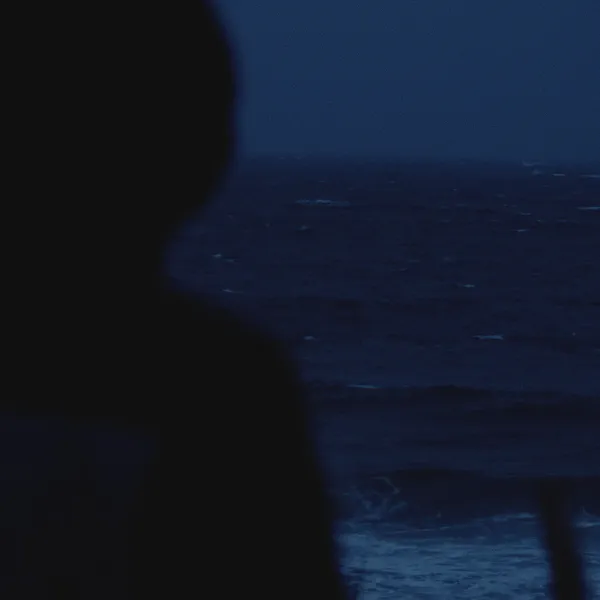 Silhouette d’une personne encapuchonnée face à une mer sombre agitée, vagues se brisant sur une jetée.