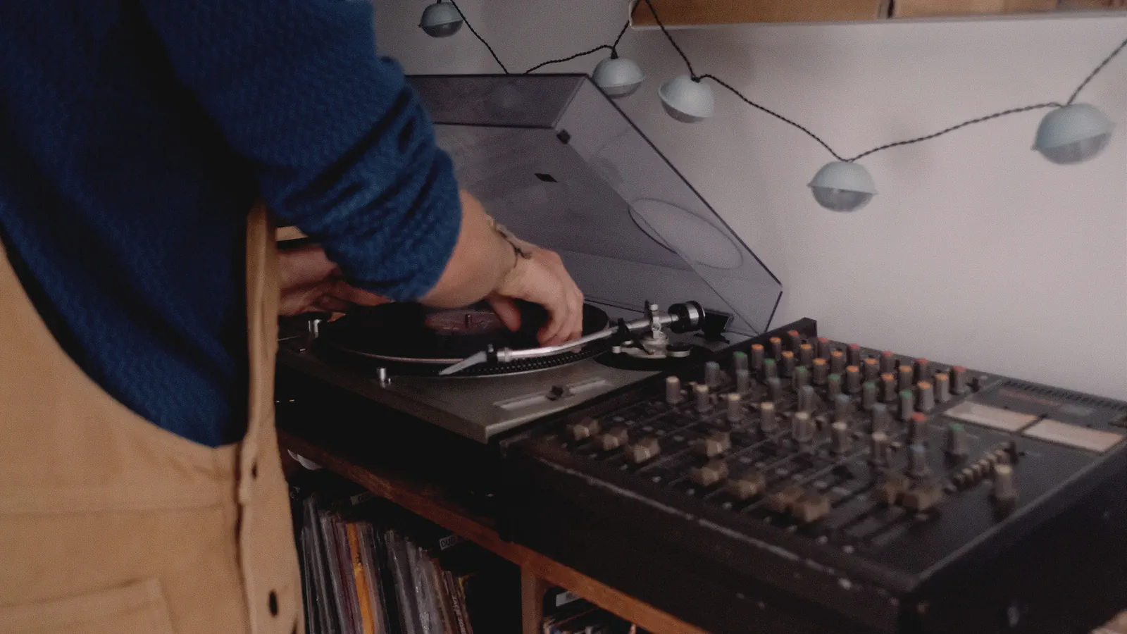 Des mains règlent la platine vinyle d’une console de mixage, guirlande lumineuse et disques rangés en dessous.