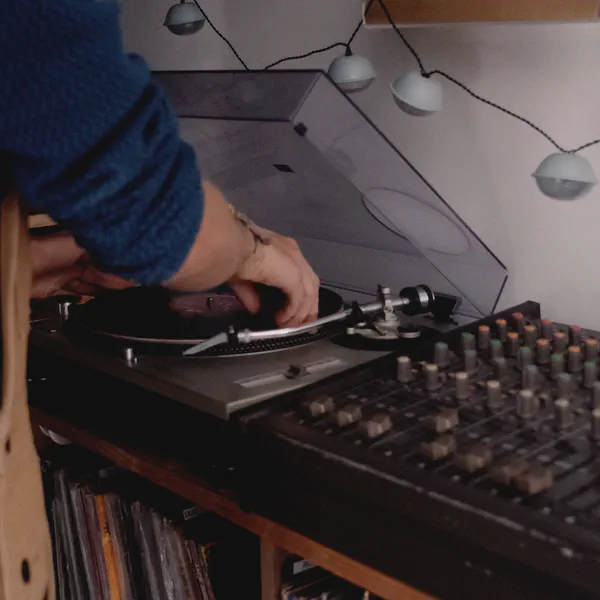 Des mains règlent la platine vinyle d’une console de mixage, guirlande lumineuse et disques rangés en dessous.