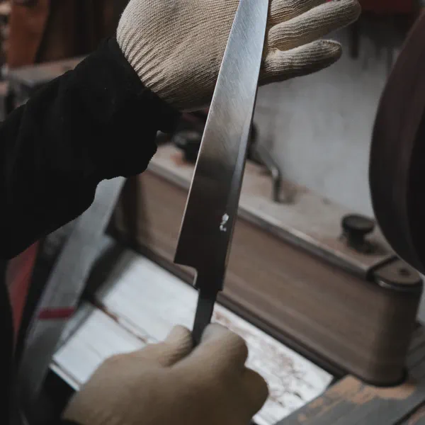 Une main gantée teste le tranchant d’une lame du doigt, outils d’atelier flous en arrière-plan.
