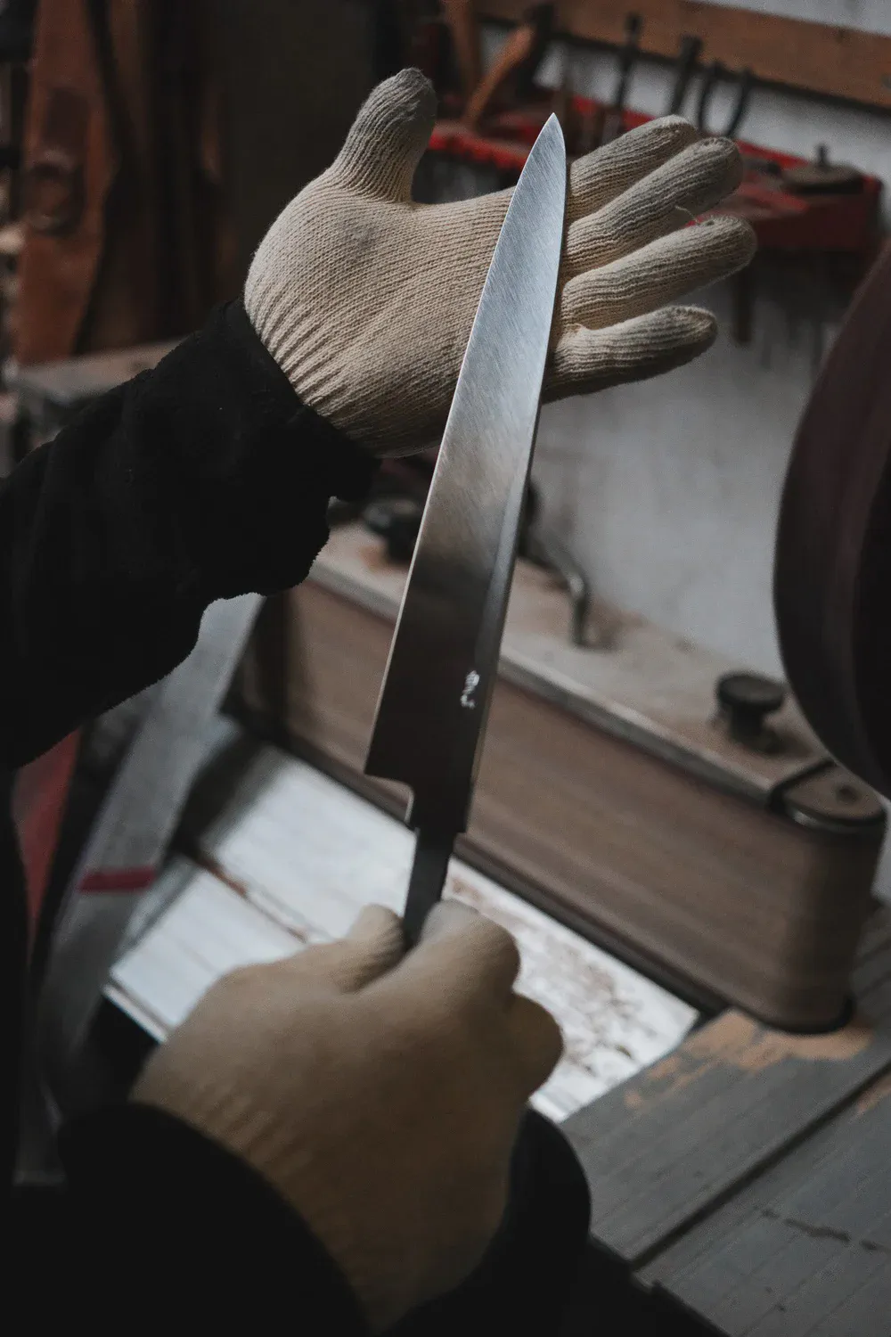 Une main gantée teste le tranchant d’une lame du doigt, outils d’atelier flous en arrière-plan.