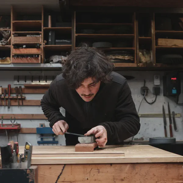 Takumi Salaün façonne un petit manche en bois avec un outil, penché sur son établi.