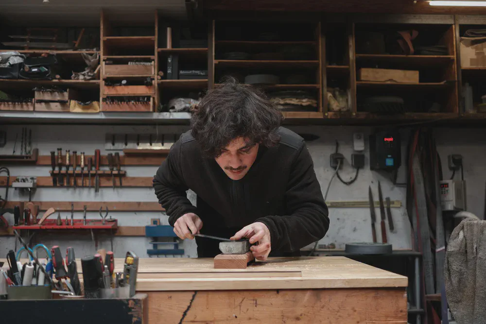 Takumi Salaün façonne un petit manche en bois avec un outil, penché sur son établi.