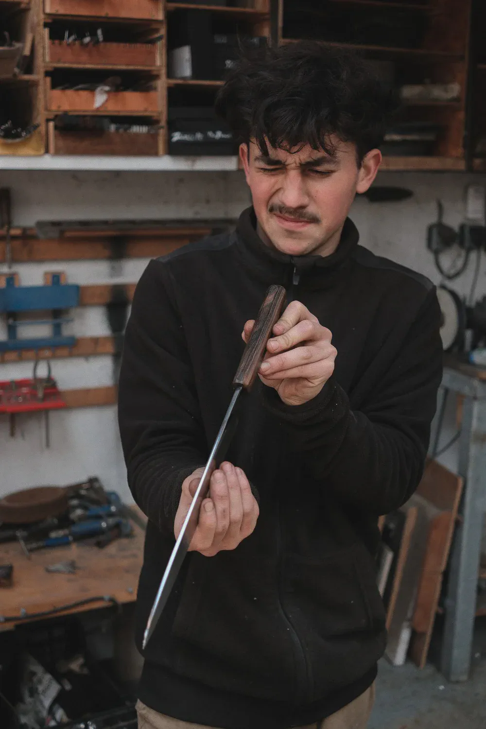 Takumi Salaün examine un couteau au manche en bois, les yeux plissés, étagères d’atelier en arrière-plan.