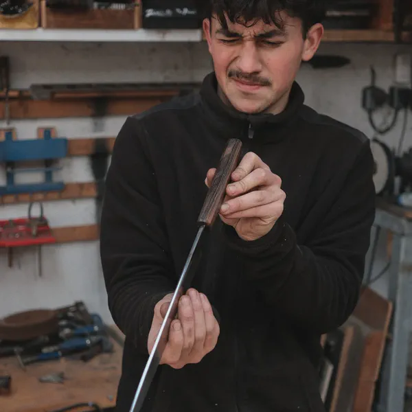 Takumi Salaün examine un couteau au manche en bois, les yeux plissés, étagères d’atelier en arrière-plan.