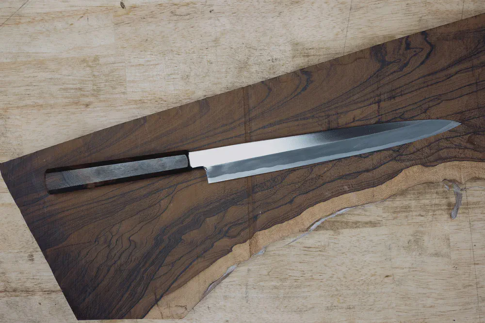 Un couteau fini au manche sombre repose en diagonale sur une planche de bois aux veines marquées.