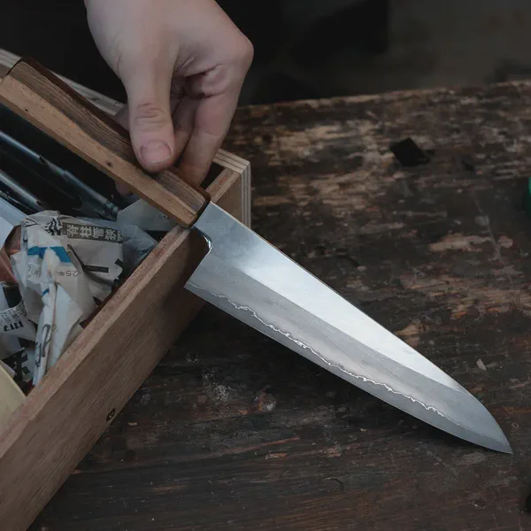 Une main tire un couteau d’une boîte en bois remplie de papiers, sur un établi usé.