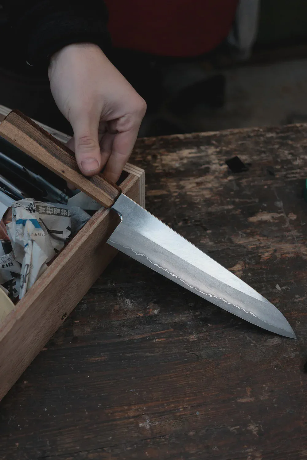 Une main tire un couteau d’une boîte en bois remplie de papiers, sur un établi usé.