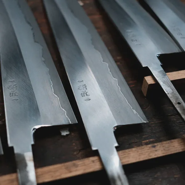 Cinq lames de couteaux alignées sur un support en bois, signature japonaise gravée près du manche.