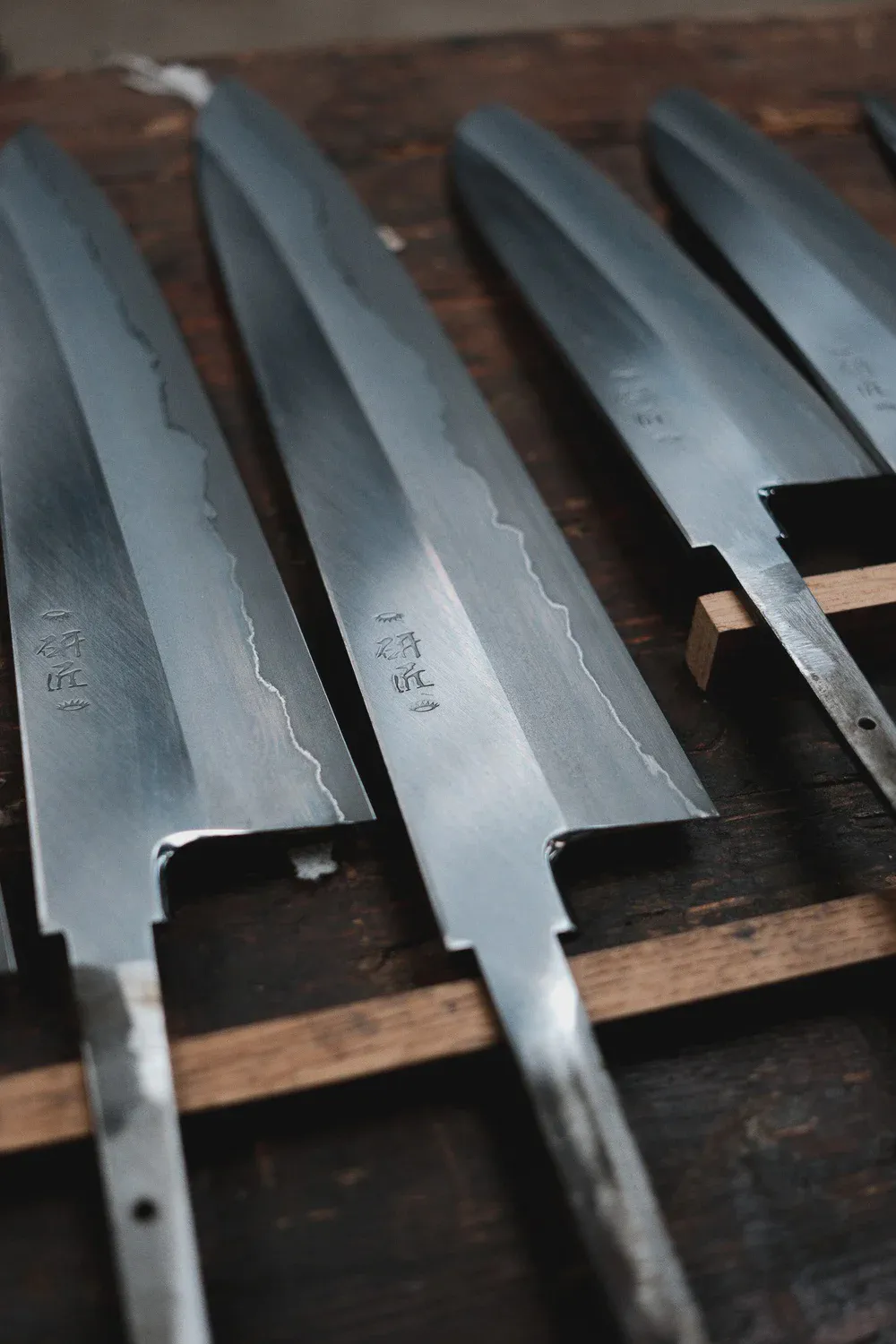 Cinq lames de couteaux alignées sur un support en bois, signature japonaise gravée près du manche.
