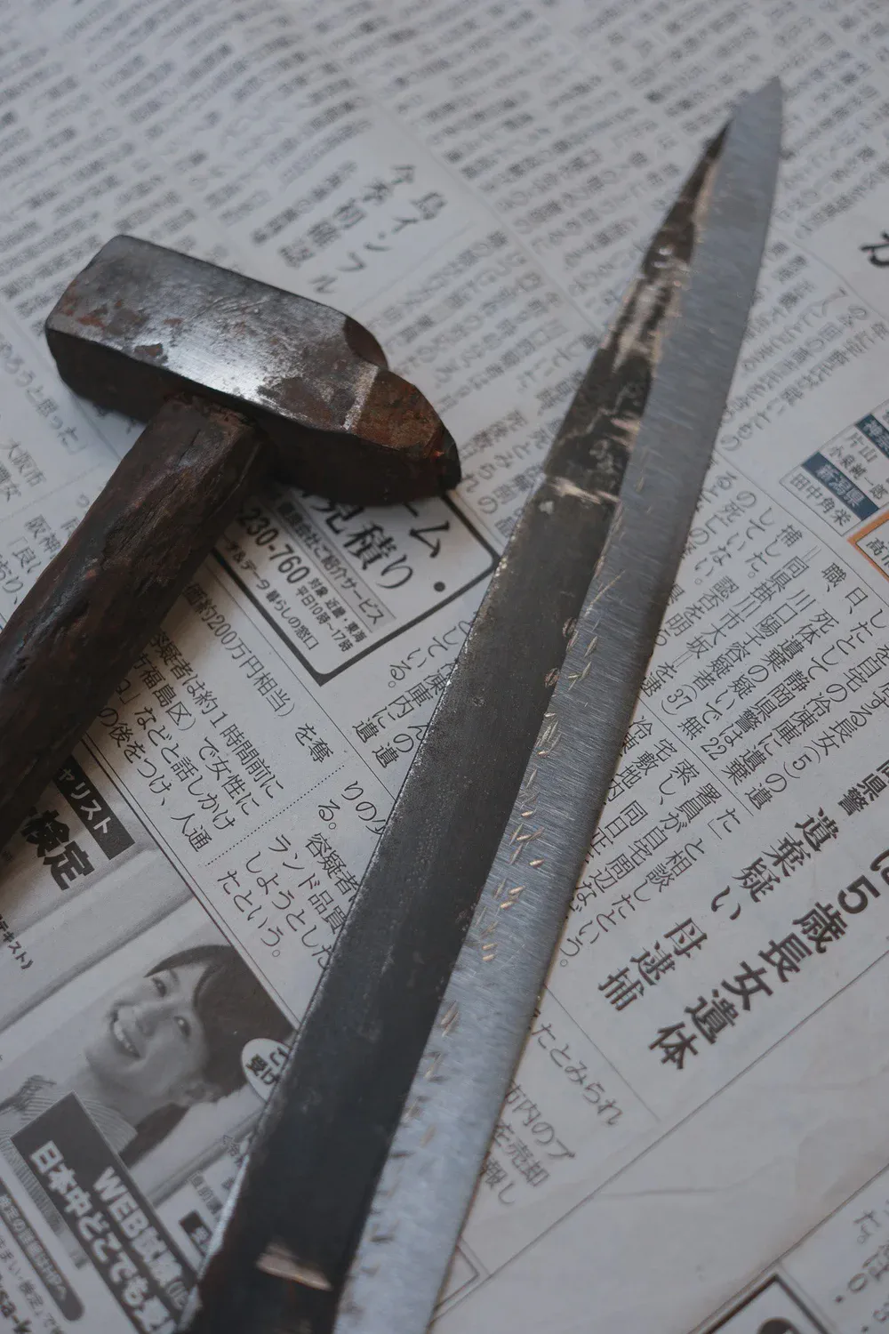 Un vieux marteau et une longue lame brute reposent sur des pages de journal japonais.