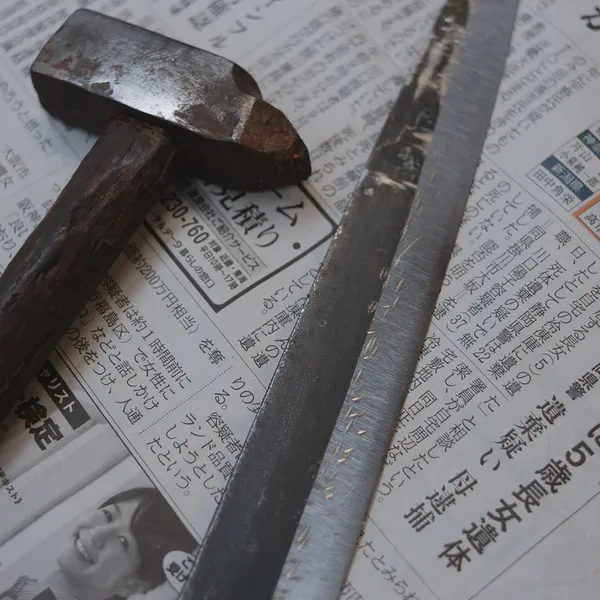 Un vieux marteau et une longue lame brute reposent sur des pages de journal japonais.