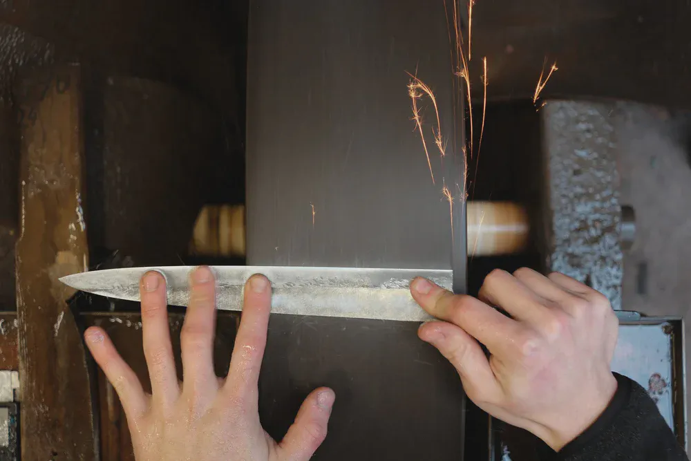 Des mains tiennent une lame contre une bande abrasive qui projette des étincelles.