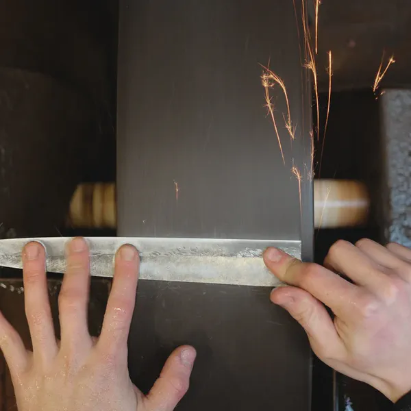 Des mains tiennent une lame contre une bande abrasive qui projette des étincelles.