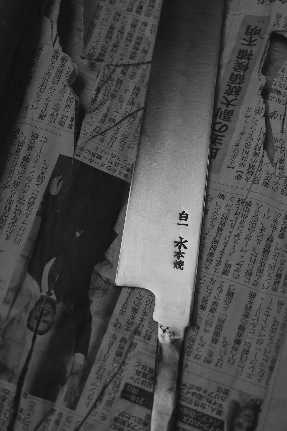 Gros plan en noir et blanc sur une lame gravée de kanjis japonais, posée sur du papier journal.