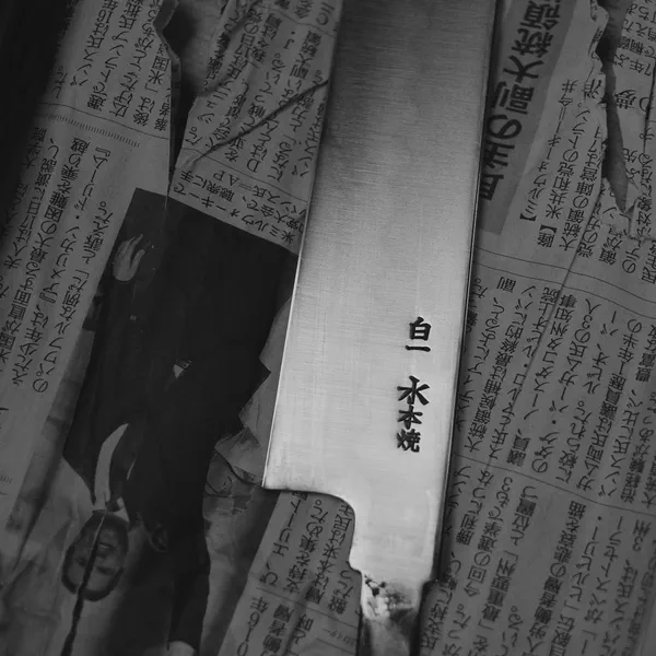 Gros plan en noir et blanc sur une lame gravée de kanjis japonais, posée sur du papier journal.