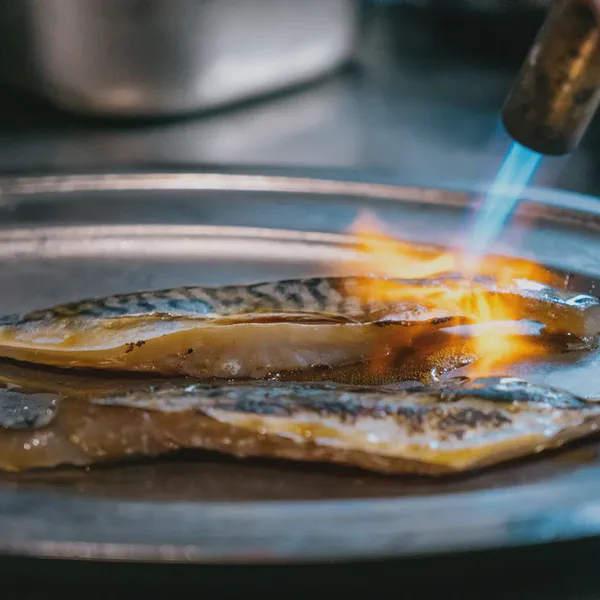 Un chalumeau grille la peau de deux filets de maquereau posés sur un plateau métallique.