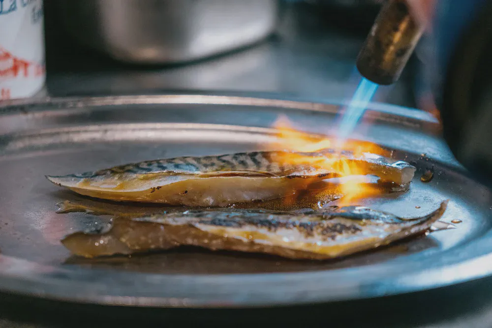 Un chalumeau grille la peau de deux filets de maquereau posés sur un plateau métallique.