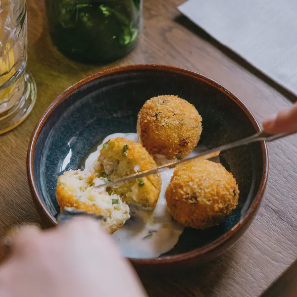 Une main utilise une pince pour servir des croquettes panées nappées de sauce dans un bol, table en bois.
