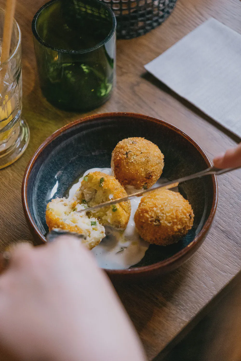 Une main utilise une pince pour servir des croquettes panées nappées de sauce dans un bol, table en bois.