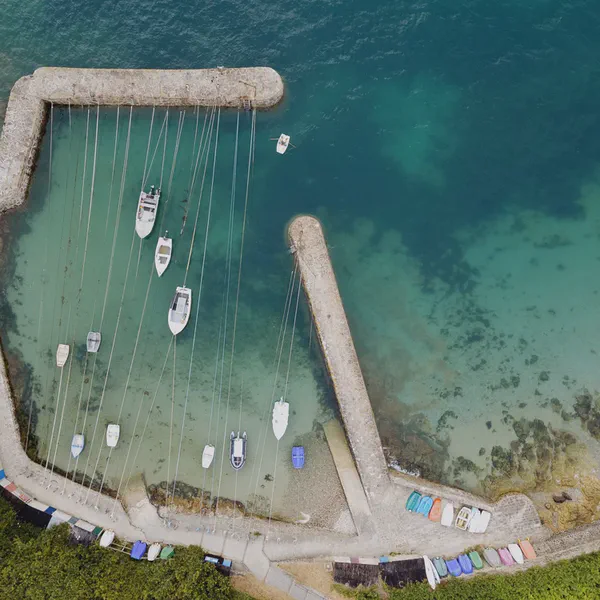 Vue aérienne d’un petit port en pierre où des voiliers sont amarrés sur des lignes, eau turquoise et annexes sur la rive.