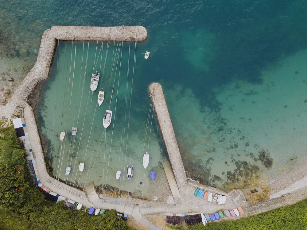 Vue aérienne d’un petit port en pierre où des voiliers sont amarrés sur des lignes, eau turquoise et annexes sur la rive.