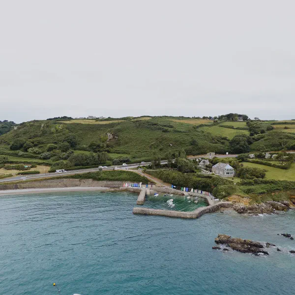 Vue aérienne d’une crique portuaire entourée de collines vertes, cabines de plage colorées près de l’eau.