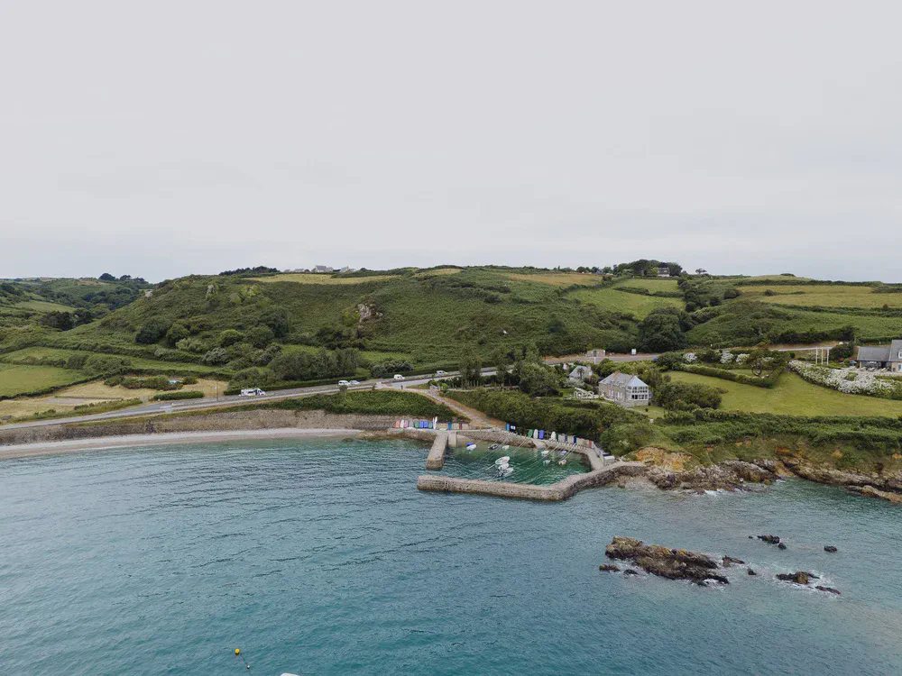 Vue aérienne d’une crique portuaire entourée de collines vertes, cabines de plage colorées près de l’eau.