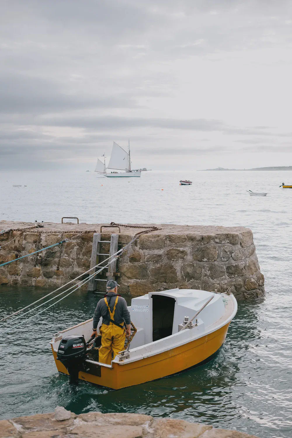 Un homme en ciré jaune se tient dans un petit bateau près d’une jetée en pierre avec échelle, voilier au loin.