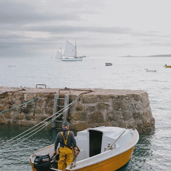 Un homme en ciré jaune se tient dans un petit bateau près d’une jetée en pierre avec échelle, voilier au loin.
