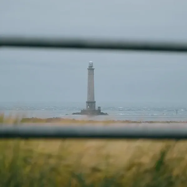 Un phare se dresse sur des rochers à marée basse, vu à travers une balustrade métallique et des herbes de dune.