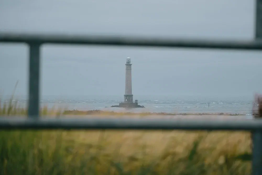 Un phare se dresse sur des rochers à marée basse, vu à travers une balustrade métallique et des herbes de dune.