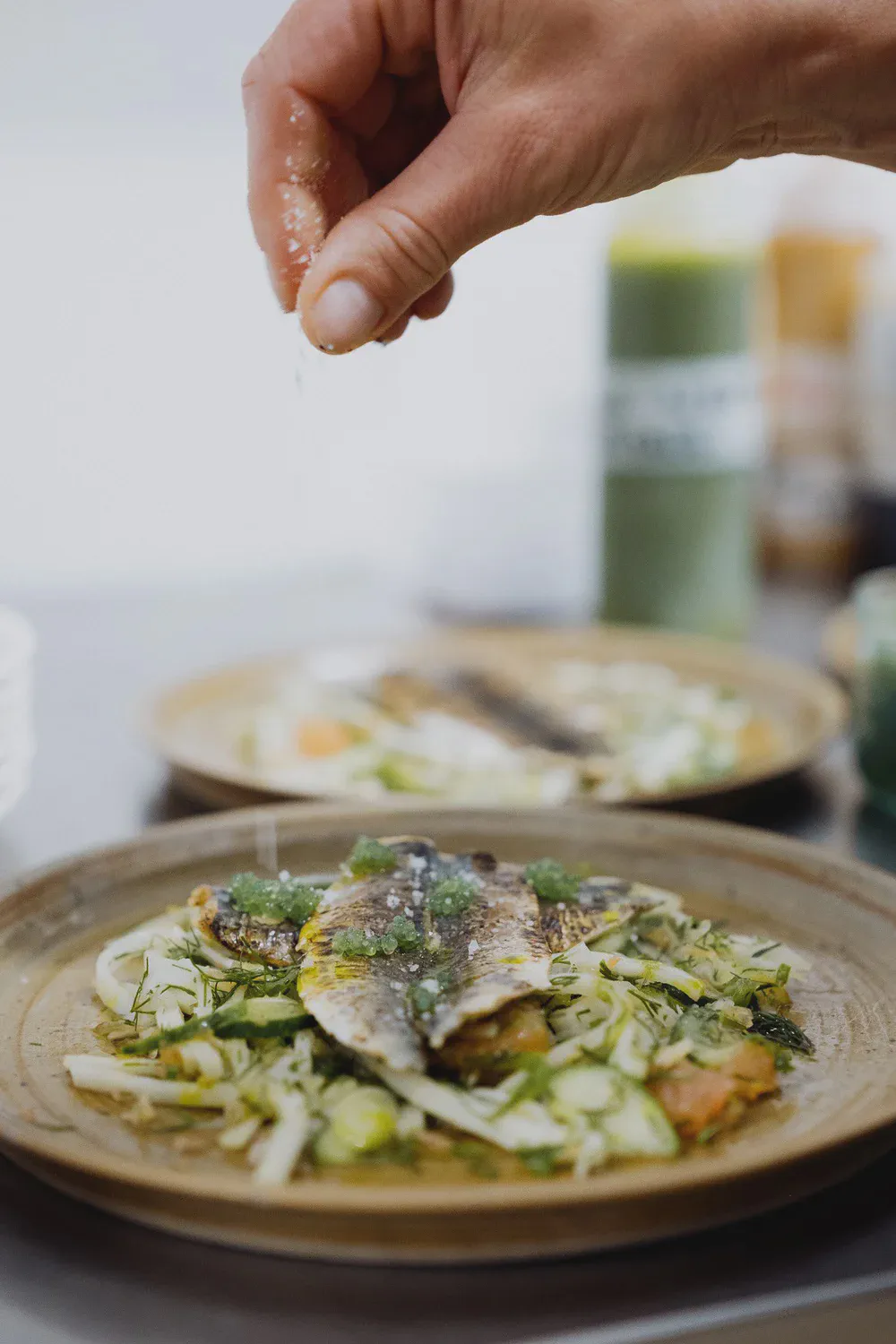 Une main saupoudre de sel un poisson grillé dressé sur un lit de légumes, second plat flou en arrière-plan.
